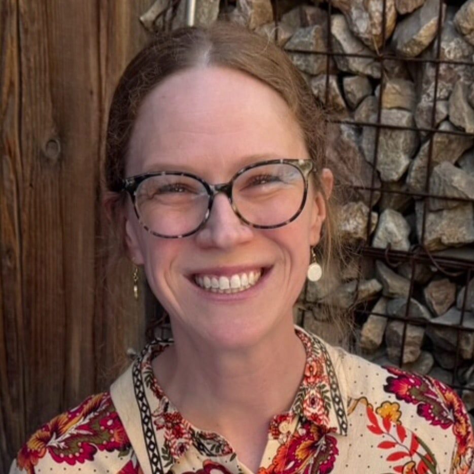 Academic Approach Tutoring and Test Prep | A woman with glasses and a floral patterned shirt smiles at the camera, sitting in front of a wooden wall and a wire basket filled with rocks.
