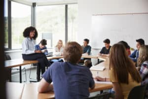 Academic Approach Tutoring and Test Prep | A teacher stands at the front of a classroom filled with diverse students seated at desks, some taking notes, in a bright room with large windows.