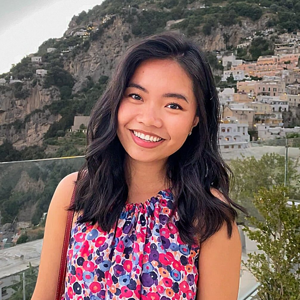 Academic Approach Tutoring and Test Prep | A woman with long dark hair wearing a colorful floral dress stands outdoors in front of a hillside town, smiling at the camera.