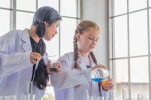 Academic Approach Tutoring and Test Prep | Two girls wearing lab coats conduct a science experiment, one pouring a blue liquid into a beaker while the other observes and holds a different container.
