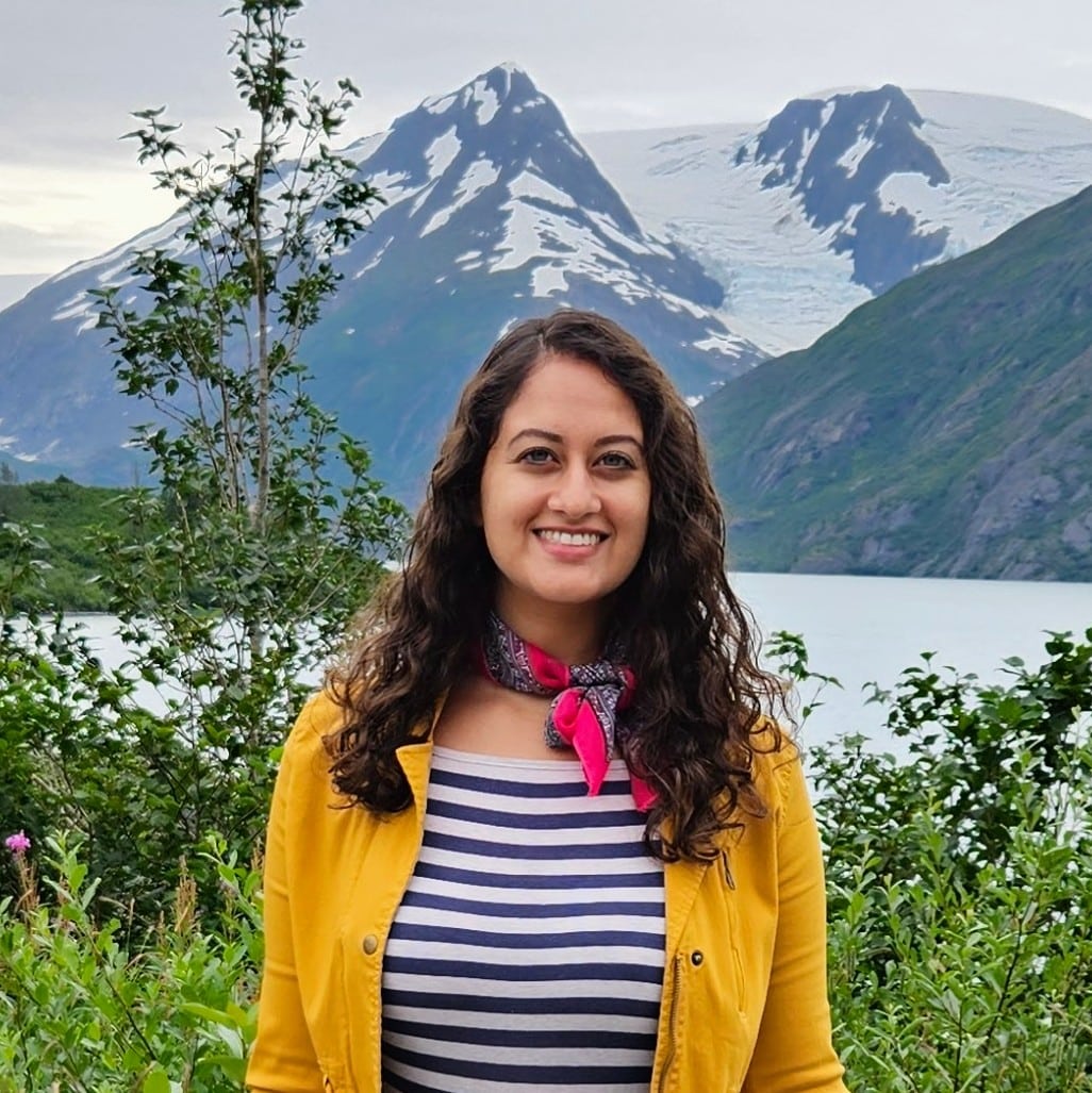Academic Approach Tutoring and Test Prep | A woman with long curly hair wearing a yellow jacket and striped shirt stands in front of a lake with snow-capped mountains in the background.