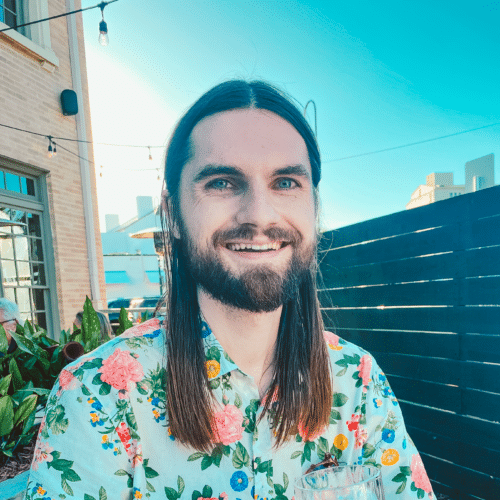 Academic Approach Tutoring and Test Prep | A smiling man with long brown hair and a beard wears a colorful floral shirt while sitting outside at a restaurant on a sunny day. There are string lights, plants, and modern buildings in the background.