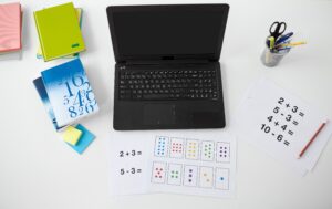 Academic Approach Tutoring and Test Prep | A desk with a laptop, notebooks, math worksheets, colored pencils, sticky notes, and a cup holding pens and scissors; worksheets show addition and subtraction problems with numbers and colored stars.
