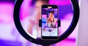 Academic Approach Tutoring and Test Prep | A smartphone mounted on a ring light records a young woman with space buns talking into a microphone, surrounded by colorful lighting and objects on a table, creating video content.