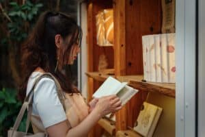 Academic Approach Tutoring and Test Prep | A woman with long hair stands beside a wooden bookshelf, reading an open book. She carries a light-colored bag and wears a white shirt with an apron. Several books and warm lights are visible on the shelves.