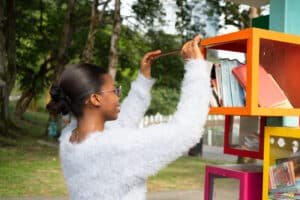 Academic Approach Tutoring and Test Prep | A woman in a white sweater and sunglasses places a book on a colorful outdoor bookshelf in a park, surrounded by trees and greenery.