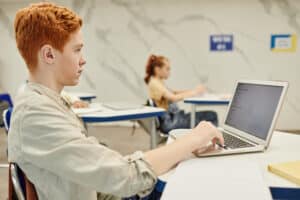 Academic Approach Tutoring and Test Prep | A red-haired student is sitting at a desk using a laptop in a classroom. Another student, slightly out of focus, is seated behind him. The classroom has a light, modern decor with minimal wall decorations.