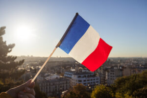 Academic Approach Tutoring and Test Prep | A hand holds a small French flag against the backdrop of a sunlit cityscape, with buildings, trees, and a clear sky visible in the background.
