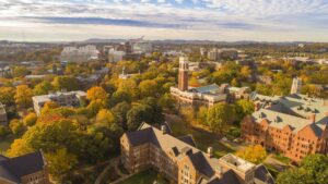Academic Approach Tutoring and Test Prep | Aerial view of a college campus with historic buildings, green and autumn-colored trees, and a city skyline in the background under a partly cloudy sky.