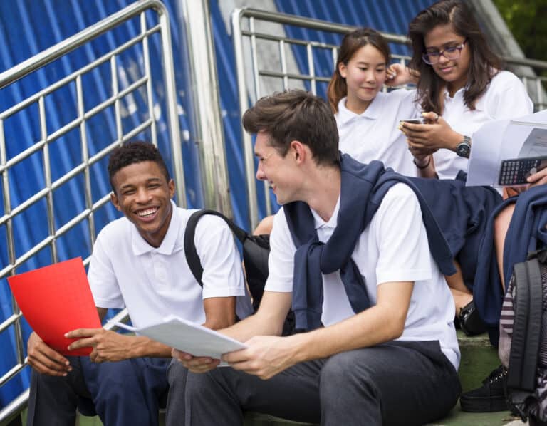 Academic Approach Tutoring and Test Prep | Five students in white polo shirts sit on outdoor steps, studying and chatting. Two are smiling with papers in hand, one checks a phone, another holds a book, and a fifth uses a calculator. A metal railing is behind them.